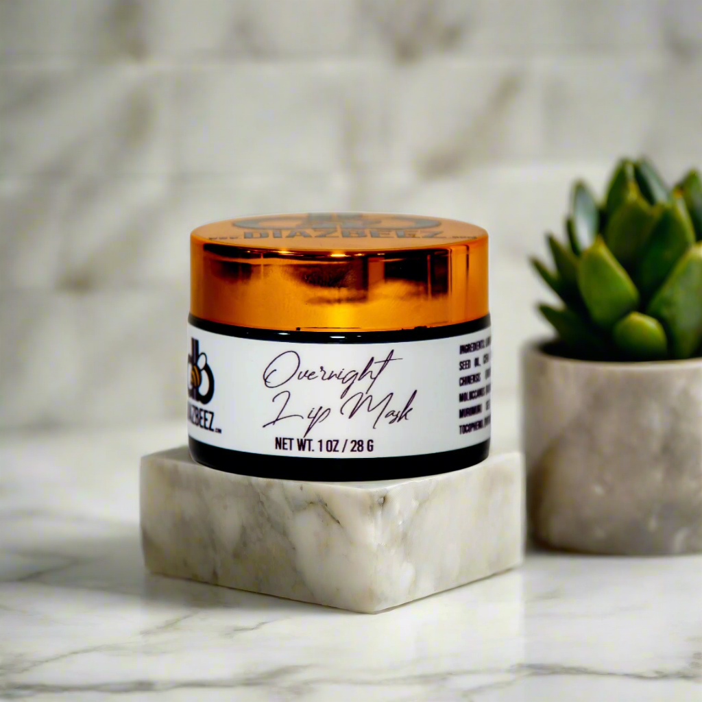 A jar of Diaz Beez's Overnight Lip Mask, featuring a distinctive orange lid, sits elegantly on a marble platform. A small succulent brings a hint of greenery to the sophisticated marble setting. Ideal for dry or chapped lips, the jar's label states "Overnight Lip Mask by Diaz Beez, Net Wt 1 oz / 28 g.