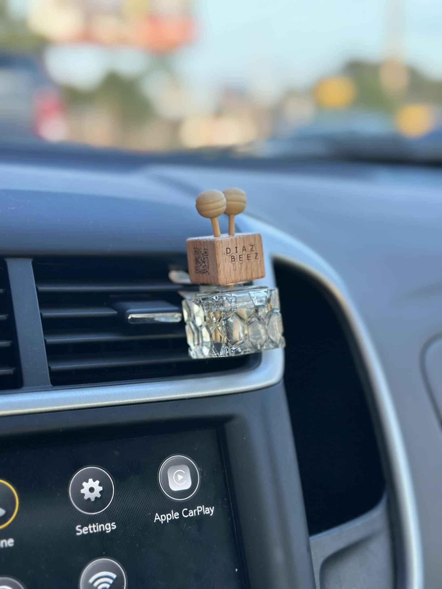 Close-up of a car air freshener attached to the vehicle's air vent. The rectangular wooden cap has two round knobs on top and reads 