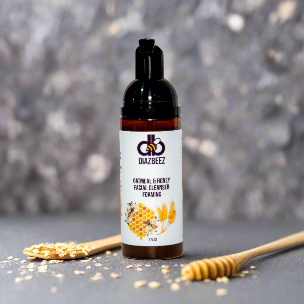 A bottle of Diaz Beez Oatmeal & Honey Foaming Facial Cleanser is centered against a blurred gray background, showcasing its natural, skin-friendly ingredients. Scattered oats accompany a honey dipper, emphasizing the gentle touch of this sensitive skin face wash.