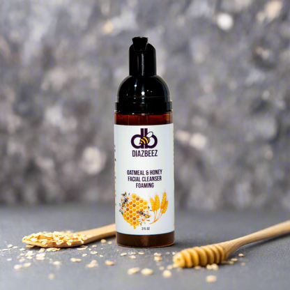 A bottle of Diaz Beez Oatmeal & Honey Foaming Facial Cleanser is centered against a blurred gray background, showcasing its natural, skin-friendly ingredients. Scattered oats accompany a honey dipper, emphasizing the gentle touch of this sensitive skin face wash.