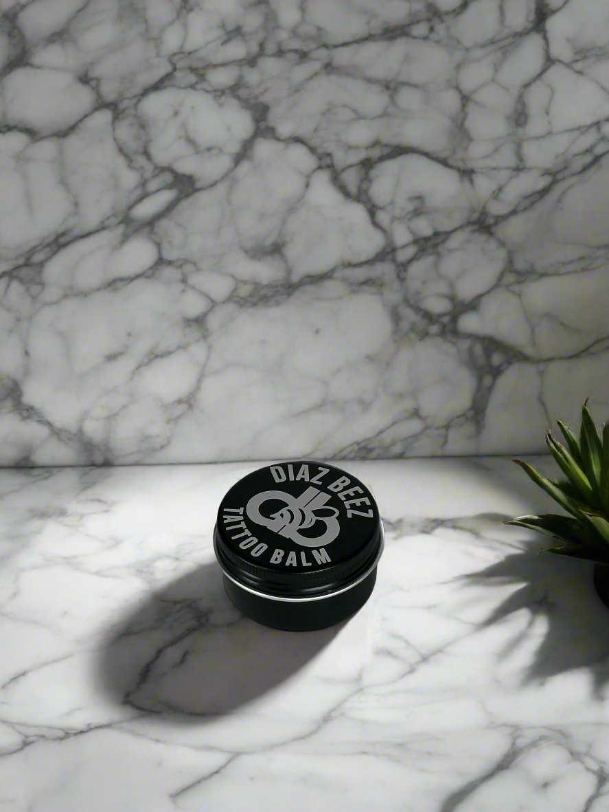 Tattoo All-Natural Balm tin on marble surface with plant, nourishing and protective for tattoo care.