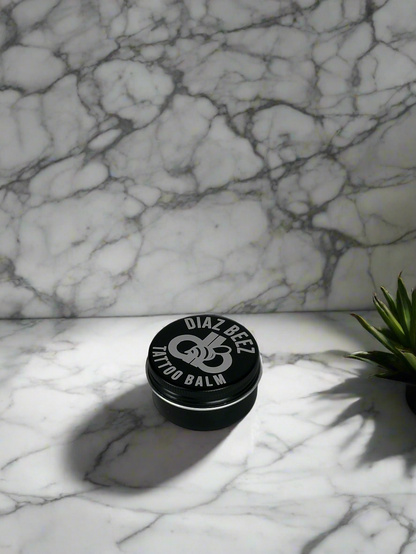 Tattoo All-Natural Balm tin on marble surface with plant, nourishing and protective for tattoo care.