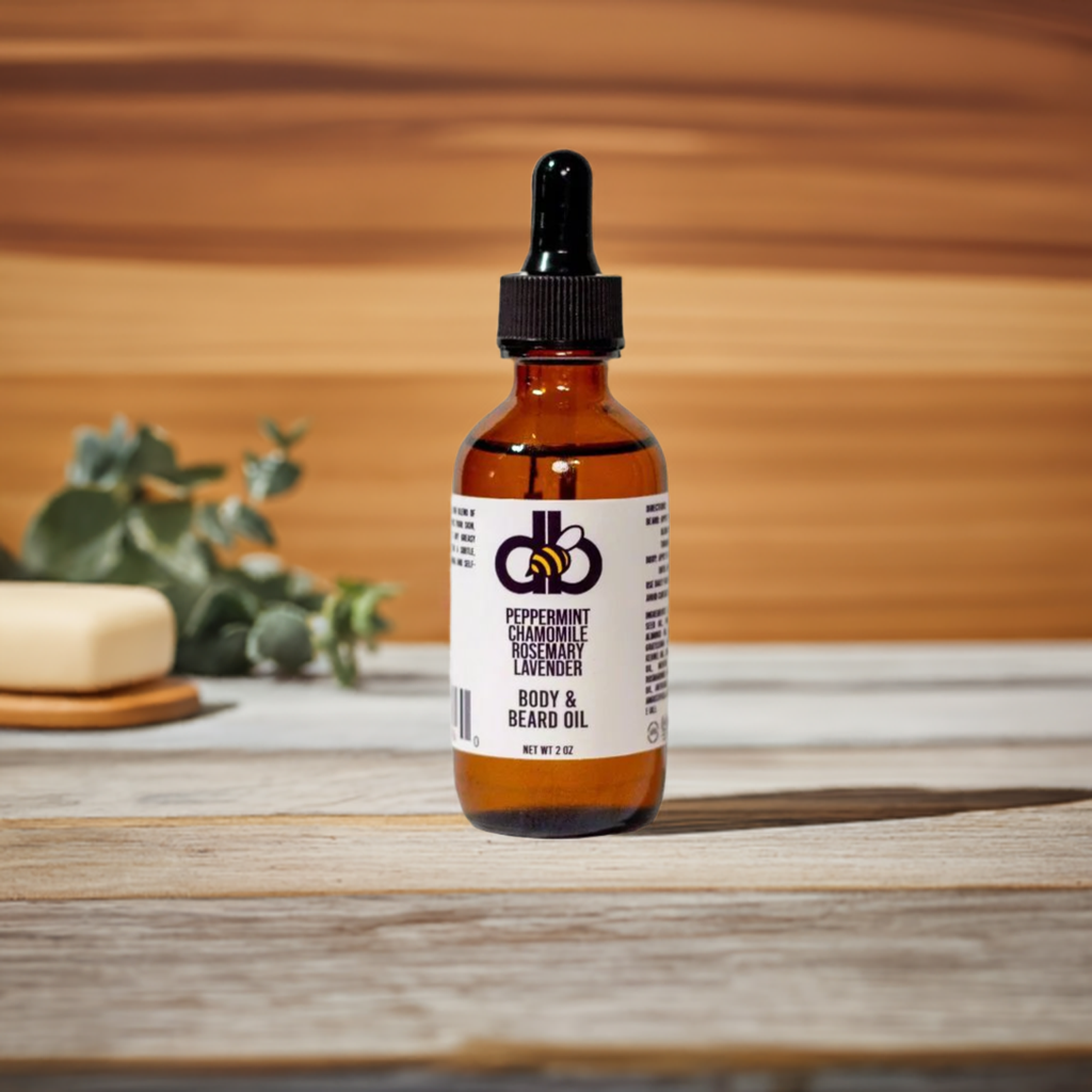 A bottle of Diaz Beez's Beard Taming Oil - Peppermint/Rosemary/Chamomile/Lavender is artfully placed on a wooden surface. In the background, a sprig of greenery and a beige soap enhance the warm, woodsy ambiance. Made with natural ingredients for an unparalleled grooming experience.