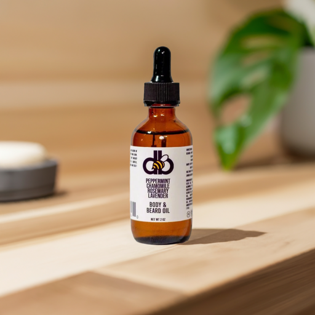 A bottle of Diaz Beez Beard Taming Oil, featuring peppermint, rosemary, chamomile, and lavender oils and crafted with natural ingredients, rests on a wooden surface. The dropper lid awaits its next use. In the softly blurred background, a potted plant and a small dish enhance the scene’s tranquility.