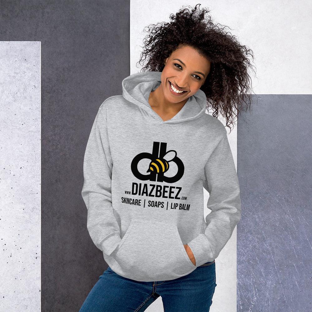 A smiling person with curly hair is sporting a cozy and stylish Unisex Hoodie from Diaz Beez, showcasing a beehive-inspired logo and the text “Diazbeez Skincare | Soaps | Lip Balm,” while standing against a geometric background of gray and purple tones.